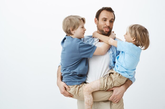 Papa liebt es, Zeit mit der Familie zu verbringen. Sorgloser glücklicher Vater, der Söhne in den Armen hält und Zunge herausstreckt