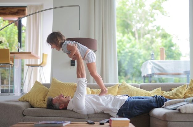 Papa liegt auf dem Sofa und hebt sein Kind im Sonnenlicht durch die Fenster in die Luft