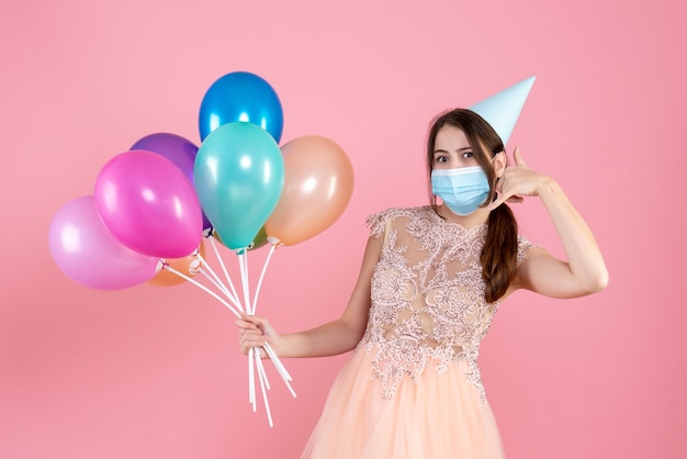 Partygirl mit Partykappe, die bunte Luftballons hält, die mich Telefongeste auf rosa anrufen