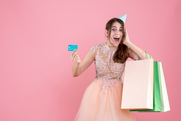Partygirl mit Partykappe, die Karte und Einkaufstaschen hält, die Hand nahe ihrer Wange auf Rosa setzen