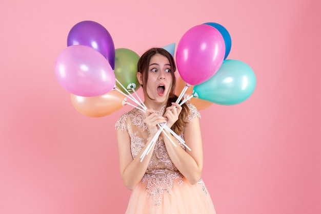 Partygirl mit partykappe, die luftballons nahe gesicht auf rosa hält