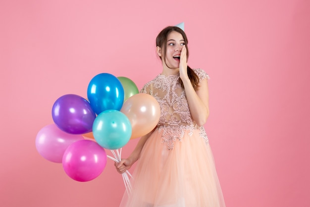 Partygirl mit Partykappe hält Luftballons, die Hand auf ihren Mund auf rosa setzen