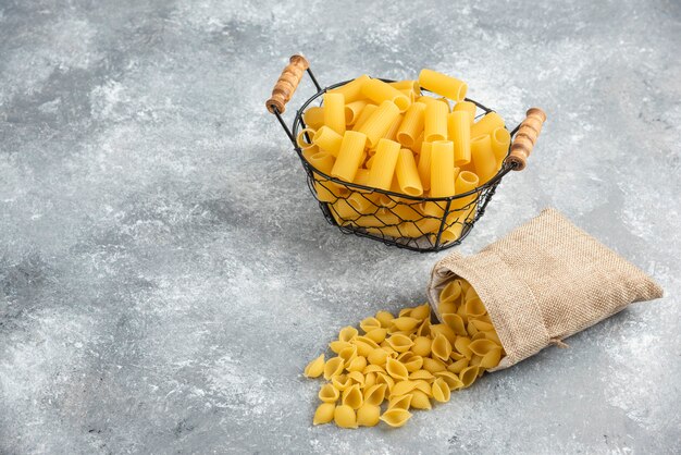 Pastasorten in einem metallischen Korb und einer rustikalen Tasche auf grauem Tisch.