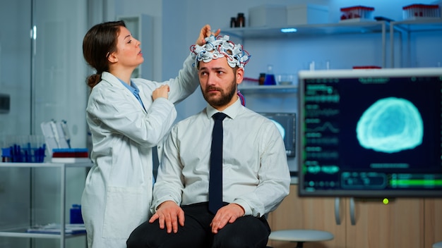 Patient mit Gehirnscan, der mit einem neurologischen Forscher diskutiert, während er das Gehirnwellen-Scanning-Headset anpasst, die Diagnose einer Krankheit untersucht, die EEG-Ergebnisse, den Gesundheitszustand und die Gehirnfunktionen erklärt