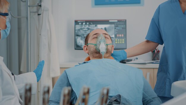 Patient mit Sauerstoffmaske, der im Zahnarztschrank eine Anästhesie erhält und sich mit zahnärztlichen Werkzeugen und Bohrern zum Polieren von Karies auf den stomatologischen Eingriff vorbereitet. Mundpflegeanästhesie beim Menschen.