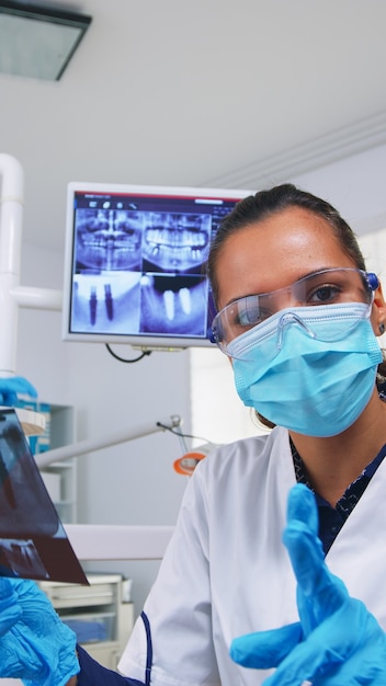 Patient pov, der den Zahnarzt anschaut und um eine zahnärztliche Röntgenaufnahme bittet, die ein Zahnbild zeigt. Facharzt für Stomatologie mit Schutzmaske, der in einer modernen stomatologischen Klinik arbeitet und die Röntgenaufnahme des Zahns erklärt
