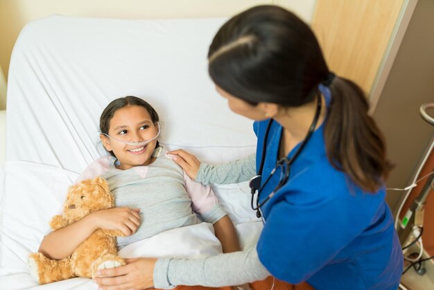 Patientin mit kleinem Mädchen, die Teddybär hält, während sie während der Behandlung dem Gesundheitspersonal auf dem Krankenhausbett zuhört