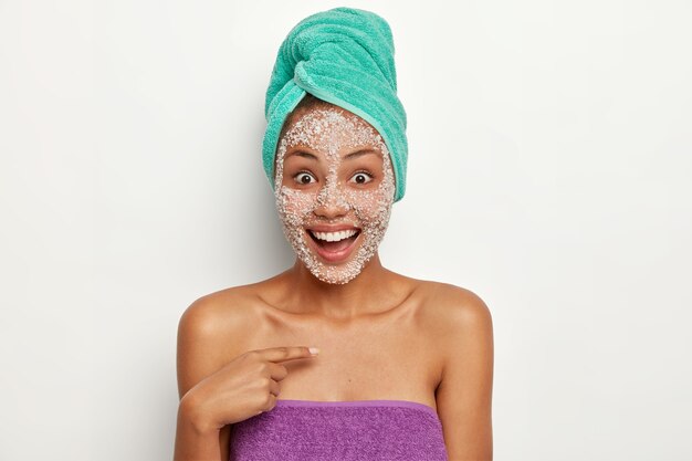 Peeling- und Reinigungskonzept. Erfreute freudige Frau zeigt auf sich selbst mit Staunen und fröhlicher Reaktion, fragt, wer ich habe, hat ein Gesichtspeeling auf der Haut, nimmt ein Bad, genießt hygienische Behandlungen