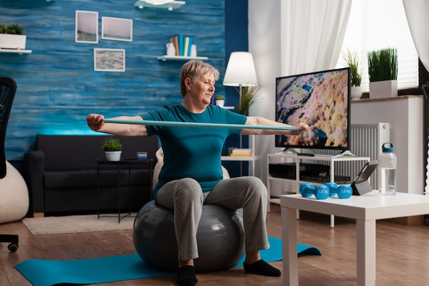 Pensionierter älterer Mann, der auf einem Fitness-Swissball im Wohnzimmer sitzt und Wellness-Fitness-Workout macht