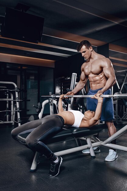 Persönlicher Trainer, der einer jungen Frau hilft, eine Langhantel zu heben, während sie in einem Fitnessstudio trainiert. Personal Trainer mit einem wunderschönen Körper ohne T-Shirt im Fitnessstudio