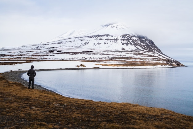 Person, die auf einem Feld steht, das durch das Meer und die Felsen bedeckt ist, die im Schnee in Island bedeckt sind