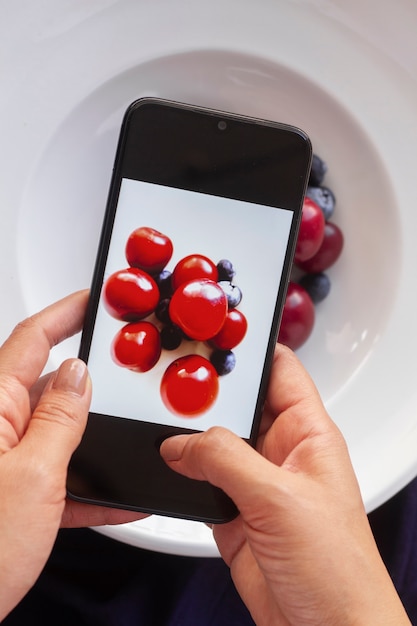 Person, die ein Foto von Kirschen und Heidelbeeren auf dem Teller mit Smartphone macht