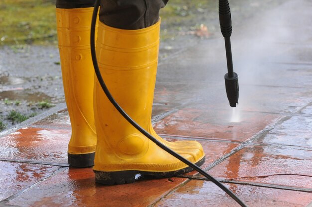 Person, die gelbe Gummistiefel mit Hochdruckwasserdüse trägt, die den Schmutz in den Fliesen reinigt