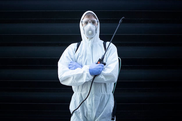 Person im weißen Chemikalienschutzanzug hält Sprühgerät mit Desinfektionschemikalien, um die Ausbreitung hoch ansteckender Viren zu stoppen