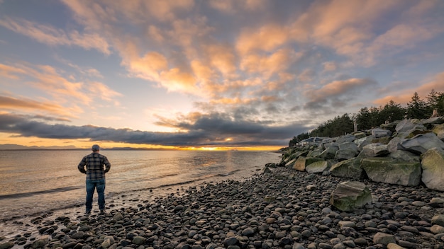 Person in der schwarzen Jacke, die auf Rocky Shore während des Sonnenuntergangs steht