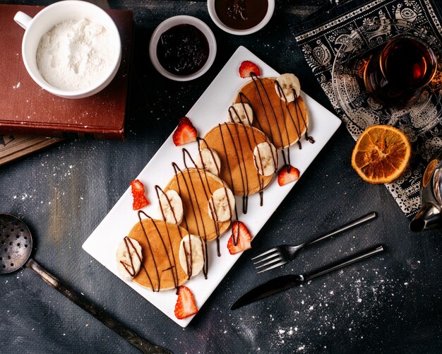 Pfannkuchen mit Draufsichten mit Bananen und Erdbeeren zusammen mit Tee und Zuckerpulver auf dem grauen Boden