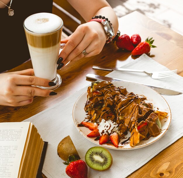 Pfannkuchen mit Seitenansicht Waffelpfannkuchen mit Erdbeer-Kiwi-Schokolade und Latte Macchiato auf dem Tisch