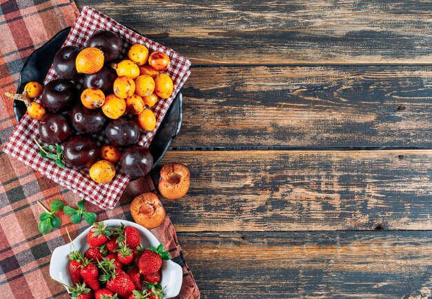 Pflaumen, Mispeln und Erdbeeren in einer Schüssel und einem Geschirrtuch mit grünen Blättern liegen flach auf einem dunklen Holz- und Picknicktuchhintergrund