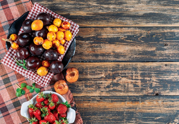 Pflaumen, Mispeln und Erdbeeren in einer Schüssel und einem Geschirrtuch mit grünen Blättern liegen flach auf einem dunklen Holz- und Picknicktuchhintergrund