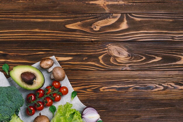 Pilz; Avocado; Kirschtomaten; Zwiebel; Brokkoli auf Tischdecke gegen Schreibtisch aus Holz