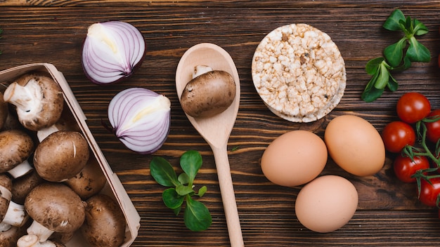 Pilz; halbierte Zwiebel; Kirschtomaten; Eier und Puffreiskuchen gegen Schreibtisch aus Holz