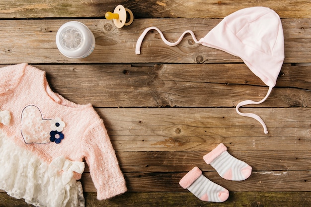 Pinkes Babykleid mit Kopfbedeckung; Paar Socken; Milchflasche und Schnuller auf Holztisch