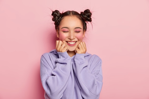 Pinup japanerin mit zwei gekämmten haarknoten berührt die wangen, hat glatte haut, trägt lebendiges rosiges make-up, piercing in der nase, trägt sweatshirt, lächelt positiv, isoliert über rosa hintergrund.