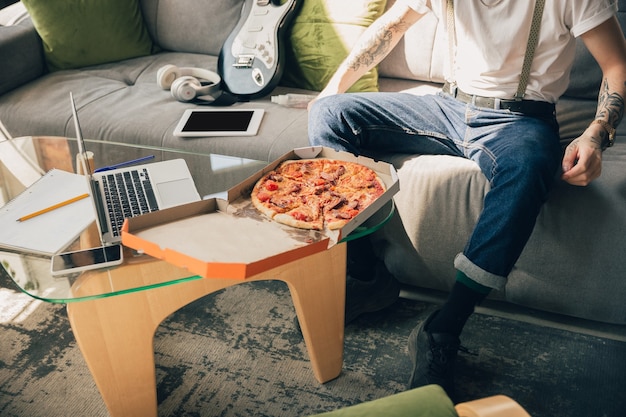 Pizza essen. Mann lernt zu Hause während Online-Kursen, intelligente Schule. Unterricht oder Beruf in der Isolation erhalten, Quarantäne gegen die Ausbreitung des Coronavirus. Mit Laptop, Smartphone, Kopfhörer.