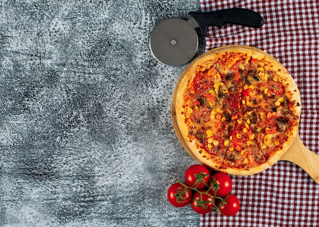 Pizza in einem Schneidebrett mit Tomaten, Pizzaschneider Draufsicht auf einen grauen Stuck und Picknicktuchhintergrund