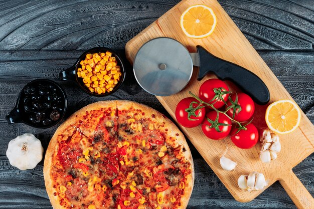 Pizza mit Knoblauch, Tomaten, einer Zitrone, Oliven, Mais und einer Pizzaschneider-Draufsicht auf einem dunklen hölzernen Hintergrund