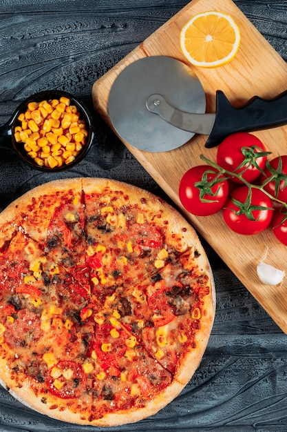 Pizza mit Tomaten, eine Scheibe Zitrone und Knoblauch, Mais und eine Pizzaschneider-Hochwinkelansicht auf einem dunklen hölzernen Hintergrund