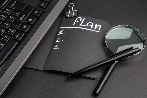 Plan von unten auf schwarzem Notizblock geschrieben Lupa Tastatur Binder Clip Stift auf schwarzem Hintergrund