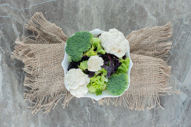 Platte mit Blumenkohl und Brokkoli auf einem mit Stoff umwickelten Sockel auf Marmor.