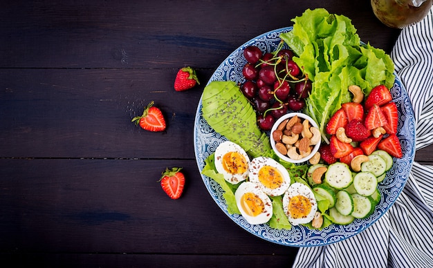 Platte mit einem Paläo-Diätfutter. Gekochte Eier, Avocado, Gurke, Nüsse, Kirsche und Erdbeeren. Paleo Frühstück. Draufsicht