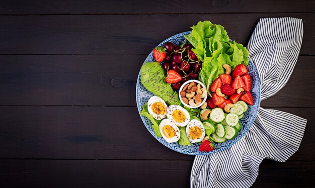 Platte mit einem Paläo-Diätfutter. Gekochte Eier, Avocado, Gurke, Nüsse, Kirsche und Erdbeeren. Paleo Frühstück. Draufsicht