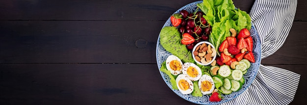 Platte mit einem Paläo-Diätfutter. Gekochte Eier, Avocado, Gurke, Nüsse, Kirsche und Erdbeeren. Paleo Frühstück. Draufsicht