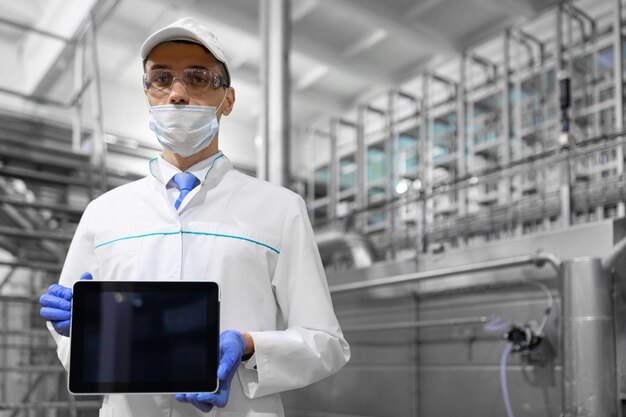 Platz zum Schreiben Der Technologe in Maskenhandschuhen und weißem Gewand steht mit einem Tablet mit leerem Bildschirm in der Fabrik Ein Mann mit einem digitalen Tablet in Uniform in der Produktionshalle