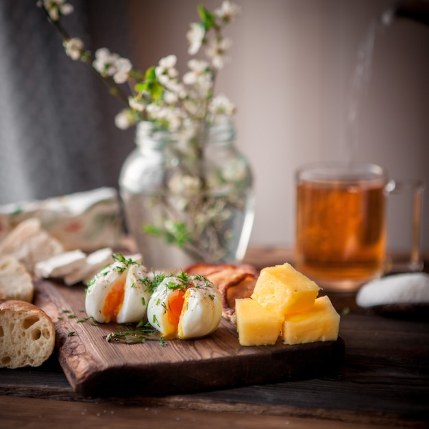 Pochiertes Ei der Seitenansicht mit Tasse Tee und Käse und Blumen im Glas im Bordkochgeschirr auf Holztisch