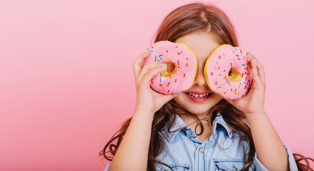 Porträt aufgeregt freudiges junges hübsches Mädchen im blauen Hemd, das Positivität ausdrückt, Spaß zur Kamera mit Donuts auf Augen lokalisiert auf rosa Hintergrund hat. Glückliche Kindheit mit leckerem Dessert. Platzieren Sie den Text