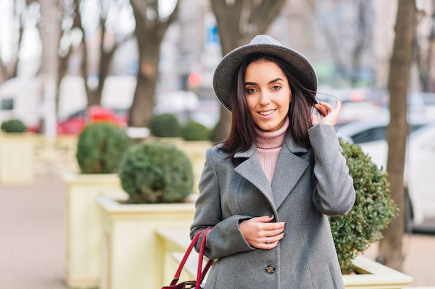 Porträt charmante junge modische Frau in grauem Hut, Mantel, der auf Straße im Stadtpark geht. Brünettes Haar, lächelnd, fröhliche Stimmung, eleganter Blick.