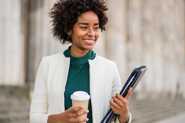 Porträt der Afro-Geschäftsfrau, die eine Tasse Kaffee und ein Klemmbrett beim Gehen im Freien auf der Straße hält. Geschäfts- und Stadtkonzept.