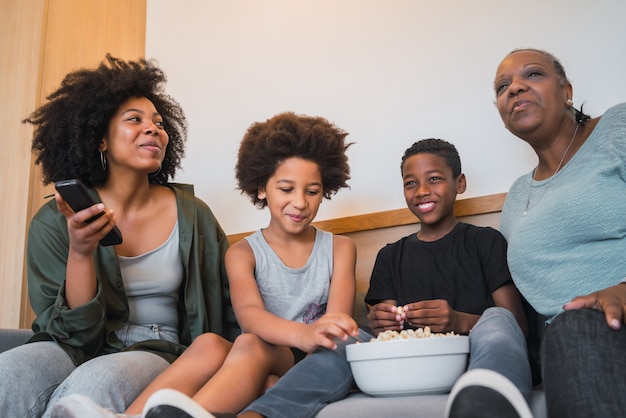 Porträt der afroamerikanischen Großmutter, der Mutter und der Kinder, die einen Film sehen und Popcorn essen, während sie zu Hause auf dem Sofa sitzen. Familien- und Lifestyle-Konzept.