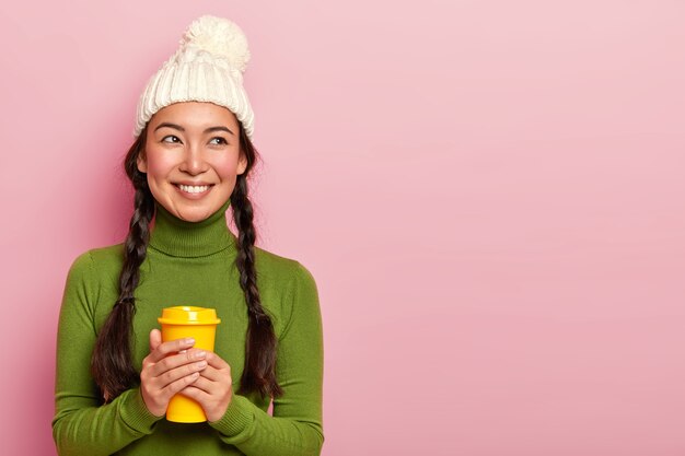 Porträt der angenehm aussehenden jungen koreanischen Frau hält Kaffeetasse, wärmt während des kalten nassen Tages, trägt weißen Hut mit Pompone und lässigem Rollkragenpullover, hat Lächeln auf Gesicht, isoliert über rosa Wand