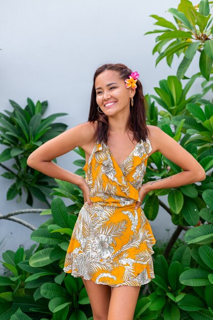 Porträt der asiatischen Frau im gelben Sommerkleid steht mit Plumeria thai Blume im Haar und in den runden Ohrringen