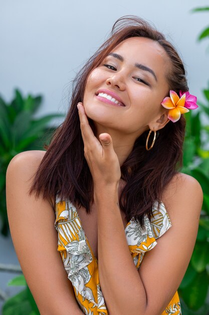 Porträt der asiatischen Frau im gelben Sommerkleid steht mit Plumeria thai Blume im Haar und in den runden Ohrringen