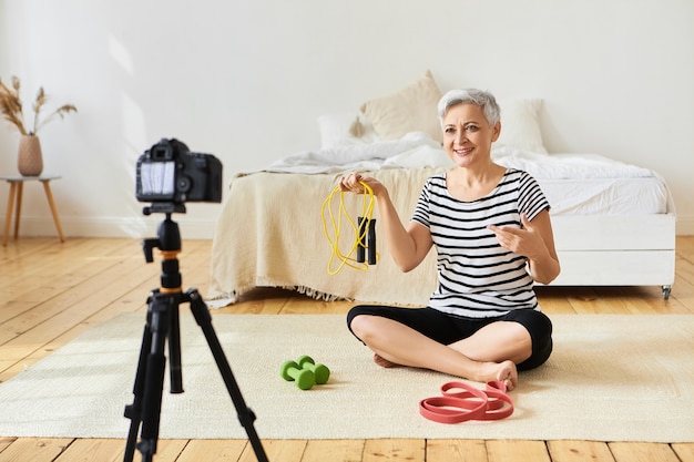 Porträt der attraktiven aktiven modernen reifen europäischen Rentnerin, die drinnen trainiert, auf dem Boden mit Sportgeräten sitzt, Springseil hält und über Cardio-Übungen vor der Kamera spricht