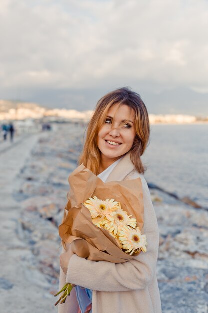 Porträt der attraktiven Frau mit Blumen, die stehen und lächeln und tagsüber liebevoll ans Meer schauen.