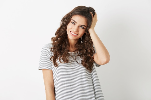 Porträt der attraktiven jungen Frau, die ihr lockiges Haar lächelnd lokalisiert auf weißem Studiohintergrund hält, gekleidet in T-Shirt, Casaualart, Blick in die Kamera, natürlicher Blick, schönes Hipster-Modell