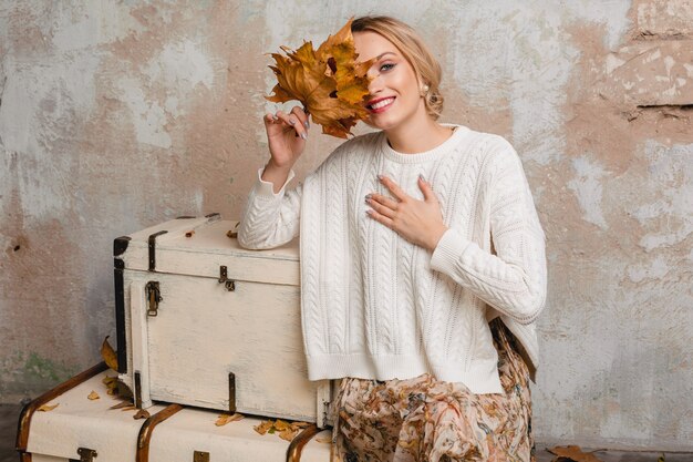 Porträt der attraktiven lächelnden stilvollen blonden Frau im weißen Strickpullover, der auf Koffern in Straße gegen Weinlesewand sitzt
