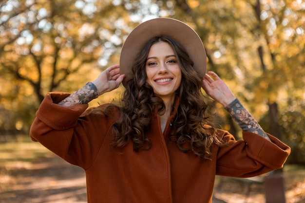 Porträt der attraktiven stilvollen lächelnden Frau mit dem langen lockigen Haar, das im Park geht, gekleidet im warmen braunen Mantel herbstlich trendige Mode, Straßenart tragenden Hut